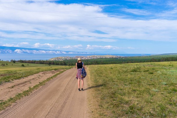 Tween tourist girl in hat and backpack walking on country road alone and admiring picturesque landscape of lake Baikal, mountains, village and blue sky. Travelling, hiking and summer vacation concept - Valokuva, kuva