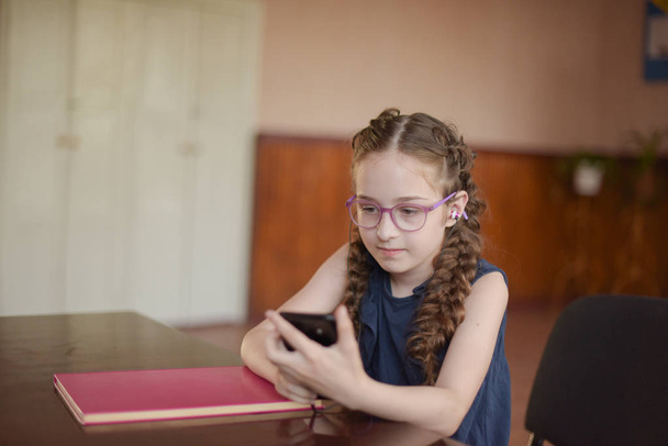 Μαθήτρια που κάθεται στο τραπέζι με σχολικά βιβλία στην τάξη. Κορίτσι σε ένα μπλε φόρεμα με κοτσίδες. - Φωτογραφία, εικόνα