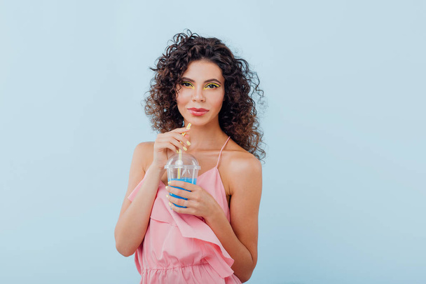 beautiful young lady with curly hair dressed in pink shirt, holding plastic cup with juice, looking at camera, isolated on blue background, copy space - Foto, Imagem