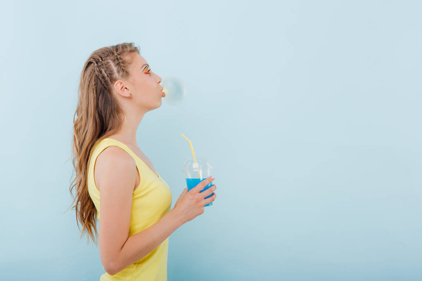 close up. young girl chewing gum bubbles, has plastic cup with juice in hand, dressed in yellow shirt, side view, positive facial emotions, isolated on blue background, copy space - Фото, изображение