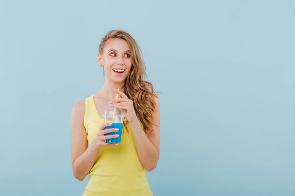 beautiful girl smiles amazed, has the plastic cup with the drink in his hand and looks sideways, dressed in yellow shirt, positive facial emotions, isolated on blue background, copy space - Photo, image
