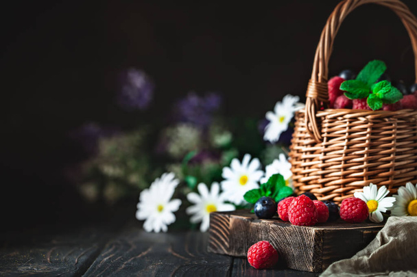 Raspberries and blueberries in a basket with chamomile and leaves on a dark background. Summer and healthy food concept. Background with copy space. Selective focus. - Fotoğraf, Görsel