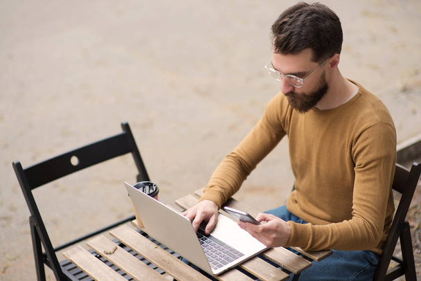 hombre barbudo guapo trabajando en el ordenador portátil y sosteniendo el teléfono inteligente en la mesa de madera
 - Foto, Imagen