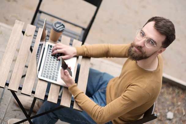knappe bebaarde man werken bij laptop en het houden van smartphone op houten tafel - Foto, afbeelding