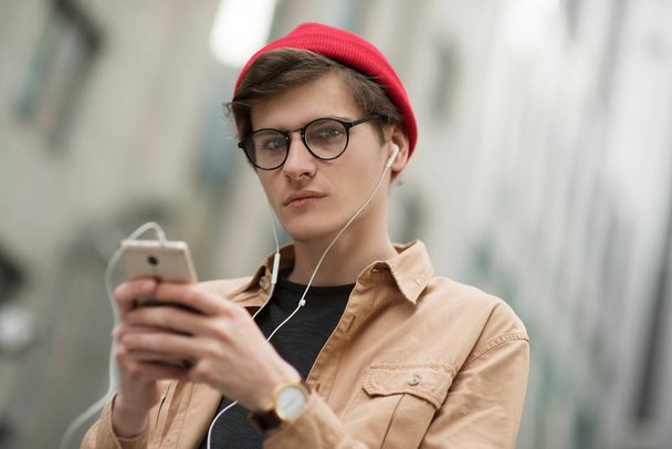 joven adolescente, un hipster, camina por la ciudad metropolitana con un sombrero rojo y una camisa elegante y mira a través de algo en su teléfono escuchando música y descansando con un buen humor
 - Foto, Imagen