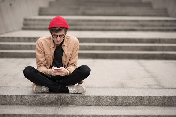 joven adolescente, un hipster, camina por la ciudad metropolitana con un sombrero rojo y una camisa elegante y mira a través de algo en su teléfono escuchando música y descansando con un buen humor
 - Foto, Imagen