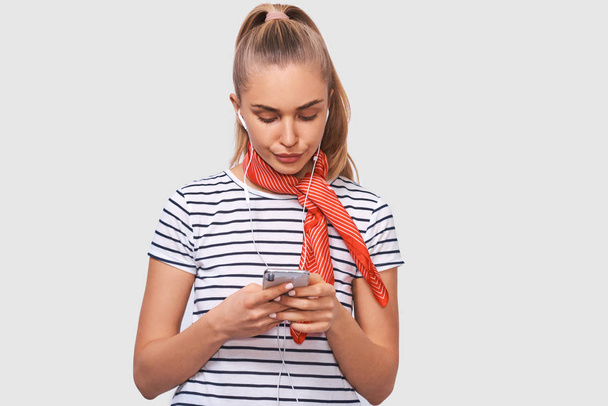 Displeased young woman using cell phone, messaging to her boyfriend. Unhappy female frowning her face and chatting with her boyfriend, looking at screen of smart phone, listening music in earphones. - Foto, Imagen