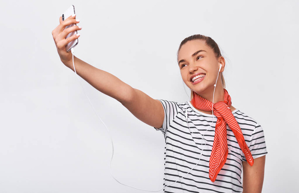 Studio horizontal portrait of beautiful European young woman taking self portrait over white wall, wearing striped t-shirt and red scarf on the neck. Happy female making a selfie on her smart phone. - Fotoğraf, Görsel