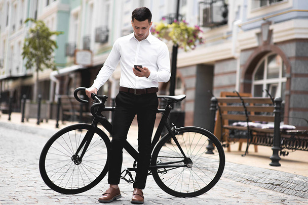 όμορφος άντρας με πουκάμισο που στέκεται με ποδήλατο και χρησιμοποιεί smartphone στην πόλη - Φωτογραφία, εικόνα