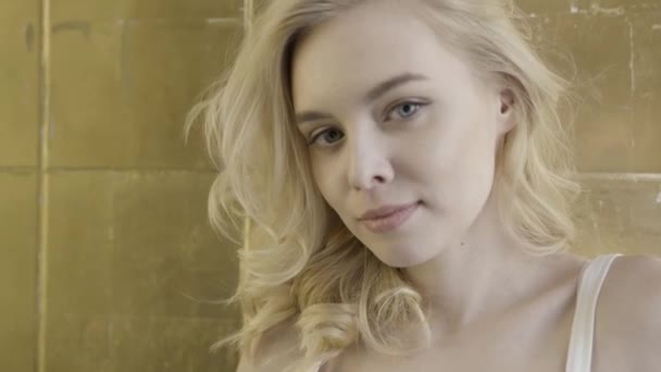 Close up for a beautiful blond girl with perceptive look over the background of golden wall tiles. Action. Young attractive woman with big blue eyes and white tank top looking straight and aside. - Footage, Video