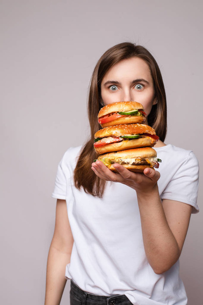 Girl with huge hamburger on hand.Studio portrait of young brunette woman in white t-shirt holding enormous burgers on her hand looking shocked or surprised at camera. - Foto, Imagem