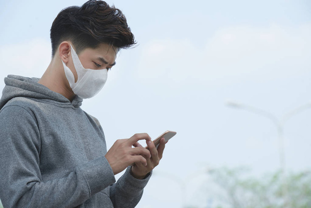 Mladý asijský muž v obličejové masce kontroluje index znečištění ve svém městě - Fotografie, Obrázek
