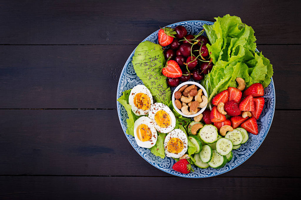 Plate with a paleo diet food. Boiled eggs, avocado, cucumber, nuts, cherry and strawberries. Paleo breakfast. Top view - Photo, Image