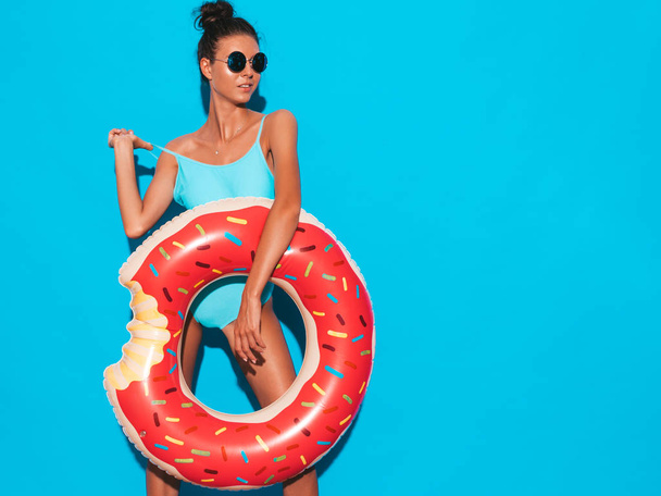 Young beautiful sexy smiling hipster woman in sunglasses.Girl in summer swimwear bathing suit with donut lilo inflatable mattress.Positive female going crazy.Posing near blue wall - Photo, image