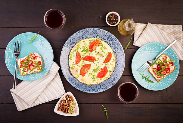 Омлет с помидорами, ветчиной, зеленым луком и сэндвичем с клубникой на тёмном столе. Фриттата - итальянский омлет. Вид сверху
 - Фото, изображение