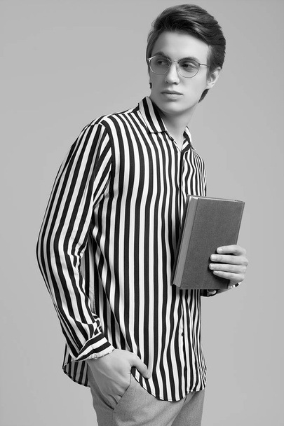 Молодой человек в полосатой рубашке позирует с книгой на сером фоне
 - Фото, изображение