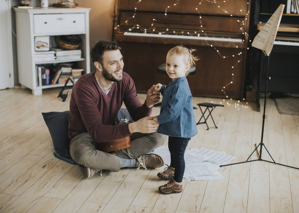 Νέος πατέρας διδάσκει τη χαριτωμένη μικρή κόρη του να παίξει κιθάρα ενώ κάθεται στο πάτωμα στο δωμάτιο - Φωτογραφία, εικόνα