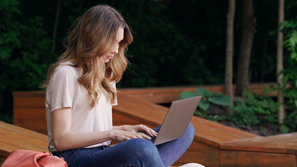 Side view of Pleased brunette woman in casual clothes using laptop computer while sitting on bench outdoors - Metraje, vídeo
