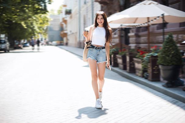 Smiley hipster meisje in een stijlvolle slijtage genieten van 's ochtends koffie te gaan terwijl het lopen van de straat op een zonnige weekend dag. Vrolijke zakelijke vrouw staande op de straat met koffie papier Cup in haar hand. - Foto, afbeelding