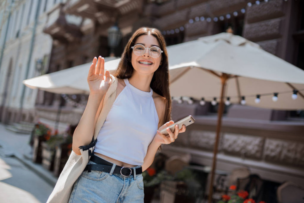 Portrait of beautiful brunette girl with phone on the street. Looking surprised, hand and smiling laughing. Urban city scene. Warm sunny weather. Outdoors walking street - Photo, Image
