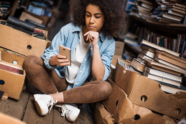 Ritratto di bella giovane donna afroamericana in camicia elegante per la correzione della visione guardando il telefono mentre aggiorna l'app al telefono e seduta vicino alla libreria in biblioteca utilizzando internet pubblico
 - Foto, immagini