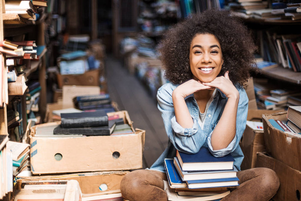 Positiivinen afrikkalainen amerikkalainen nuori nainen pukeutunut rento kulumista nauraa ja katselee pois. Iloinen tumma iho opiskelija hymyilee istuessaan työpöydällä monien opiskelu kirjoja tentti valmistelu
 - Valokuva, kuva