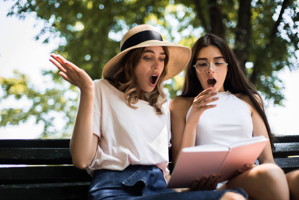 朝の公園で読書をする驚いた若い女性のショット。通りに座って本を見ている二人の女の子。学生のコンセプト - 写真・画像