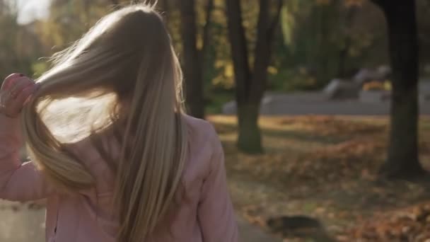 Smiling girl turns around and smiles looking at camera in autumn park in slow motion. Portrait of attractive blonde woman at sunset. Sun is shining. Medium shot - Footage, Video