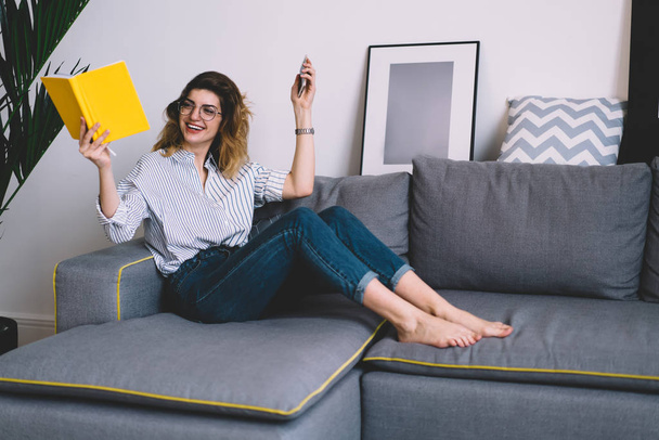 Cheerful stylish woman enjoying leisure lying on cozy couch in fashionable apartment, happy excited female reader with book and smartphone in hands rejoicing recreation time in home interior - Valokuva, kuva
