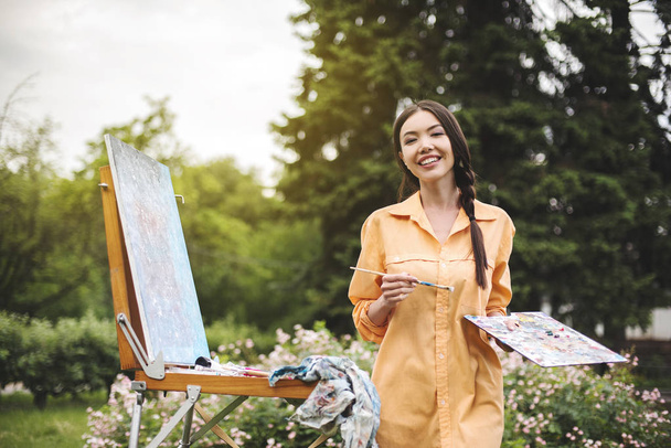 Egy fiatal nő művész rendelkezik ecsettel és fest egy képet egy festőállvány a sugarak a naplemente. A festő festékek Olajfestmények naplementekor. Blogger művész festő hogy videó órákat az otthoni kertben - Fotó, kép