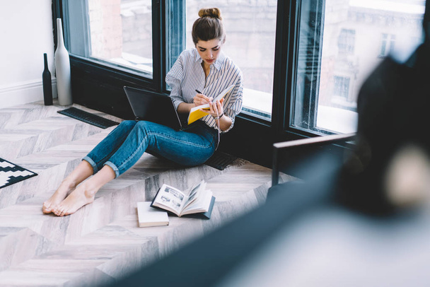 Clever student spending time for preparing to university course work using modern equipment for studying, skilled female journalist working remotely on project while sitting at floor in apartment - 写真・画像