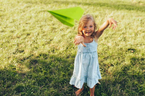 Happy pretty little girl playing with a paper plane in summer day in park. Cute kid throwing a airplane outdoors in the garden. Childhood concept - Foto, imagen