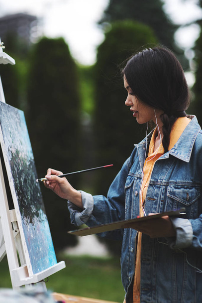 Creative pensive painter paints a colorful picture. Closeup of painting process in art workshop. Creative positive woman painter paints draw in her studio outdoor in park abstract picture of flowers. - Φωτογραφία, εικόνα