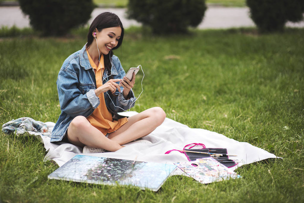 Portrait attractive brunette woman with paint art to go recreating in city park holding mobile phone, young female student looking at mobile spending break sitting on grass in college campus outdoors - Foto, Imagen