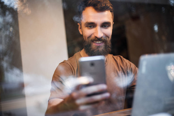 Боковой вид мужчины-блогера, просматривающего профиль в сетях на смартфоне через интернет 5G, в стильной кофейне с современным ноутбуком.
 - Фото, изображение