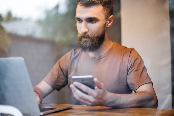 Portrait of handsome bearded caucasian male lreading online magasines artisles connected to the laptop sitting indoors on a raining day. Student guy surfing the web on laptop connected to public wifi - 写真・画像