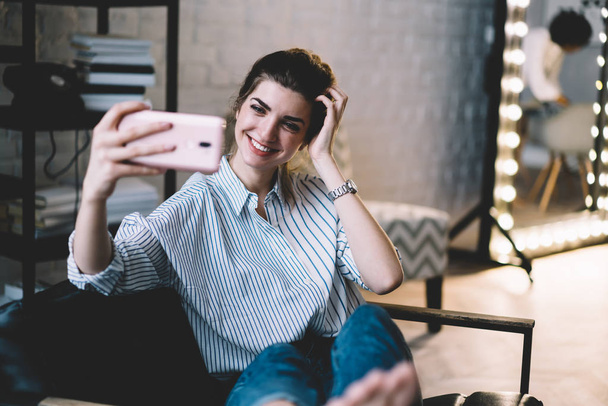 Carefree καυκάσιος hipster κορίτσι αίσθηση απόλαυση από στιγμές χαλάρωσης στο άνετο διαμέρισμα χαμογελώντας και τη λήψη φωτογραφιών μέσω της μπροστινής κάμερας στο σύγχρονο κινητό τηλέφωνο για τη διατήρηση της μνήμης της καλής στιγμής - Φωτογραφία, εικόνα
