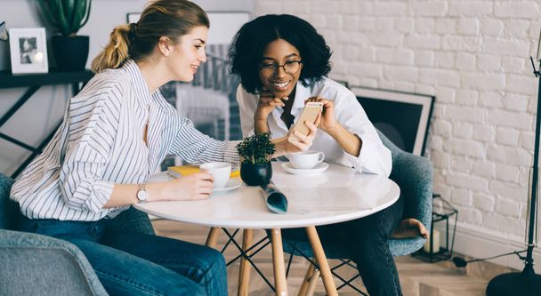Caucasian female blogger showing smartphone with new publication on website to afro american friend dressed in casual wear during positive meeting at table with tasty coffee in stylish apartment - Photo, image