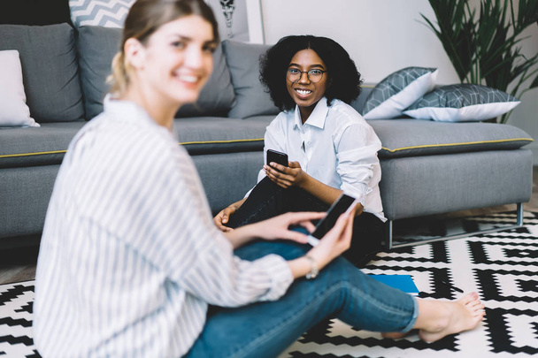 Portrait of cheerful afro american female blogger in optical spectacles looking at camera with cellular phone in hand sitting on apartment floor with millennial caucasian friend on blurred frontage - Valokuva, kuva