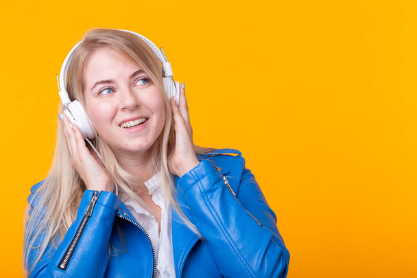 Portrait of cute positive young blonde girl listening to favorite music in headphones in blue leather jacket posing on yellow background. Concept of music concerts and online radio. Copyspace - 写真・画像