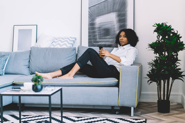 Pensive black hipster girl lying on comfortable couch in stylish apartment and thinking about notification for cellular phone during leisure time for recreation, thoughtful woman with cellphone - Φωτογραφία, εικόνα