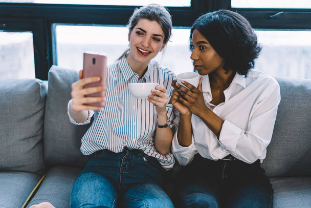 Attractive black skinned girl together with cheerful caucasian friend posing on front camera of smartphone to make cool selfie for publication in social networks, happy women taking photo on cellular - Фото, изображение