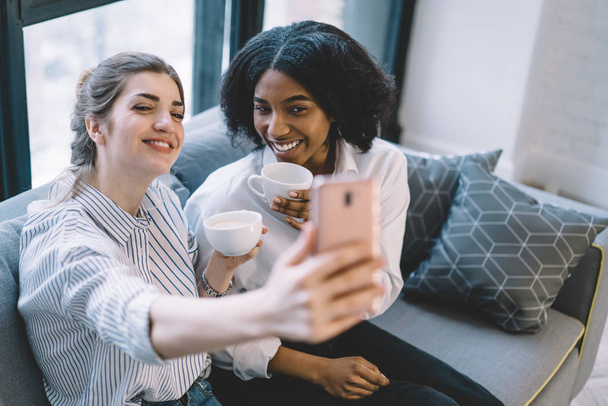 Happy two women friends smiling at front camera of digital smartphone and making photos, cheerful multicultural girls taking selfie on cellphone at home interior during conversations with coffee - Foto, Imagen