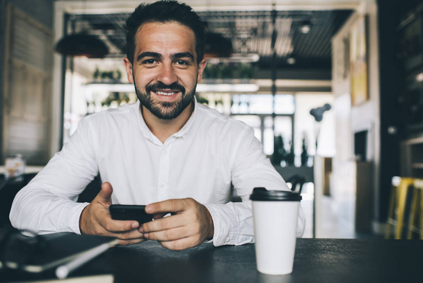 Portrait of positive successful male entrepreneur with mobile phone in hands spending work break in coffee shop sitting at cafeteria table with takeaway caffeine beverage and smiling at camera - Photo, image