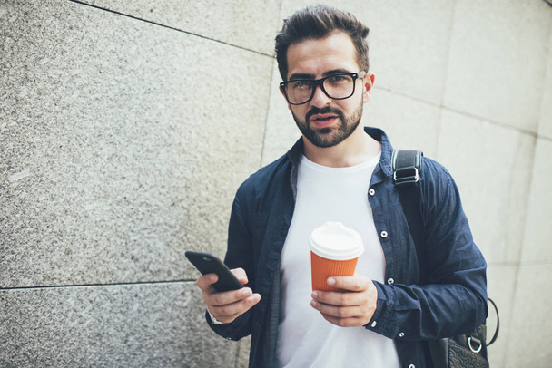 Half length portrait of millennial man with coffee to go using cellphone device for online communication, trendy dressed male blogger with modern smartphone gadget looking at camera outdoors - 写真・画像