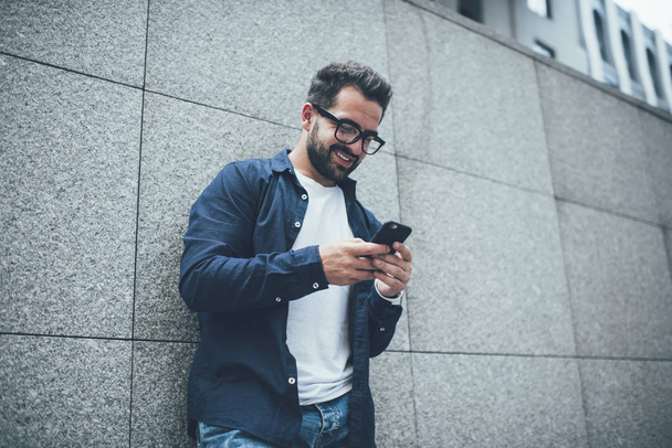 Positive male blogger in optical spectacles for vision correction standing near city wall and typing content text for sharing while networking, happy hipster guy watching funny video on website - Фото, изображение