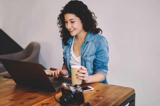 Счастливая девушка-хипстер, одетая в повседневную одежду, смотрит интересное видео на сайте через ноутбук, подключенный к Wi-Fi, веселая женщина-фрилансер, наслаждающаяся кофейным напитком во время работы в Интернете - Фото, изображение