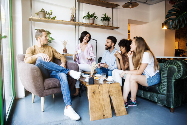 Grupo de jóvenes sentados en un café interior moderno, conversando y disfrutando juntos, tomando café y divirtiéndose
 - Foto, Imagen