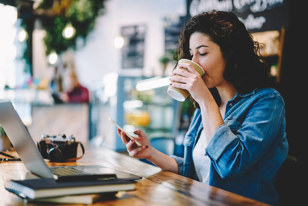 Caucasian brunette woman dressed in casual wear enjoying caffeine beverage during messaging with followers from own web blog connected to 4g wireless on smartphone device for browsing internet - Foto, Imagen