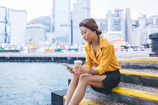 Side view of millennial female generation in trendy wear typing text for search destination way via location application on modern cellphone gadget, Asian tourist using 4g internet during coffee break - Foto, afbeelding
